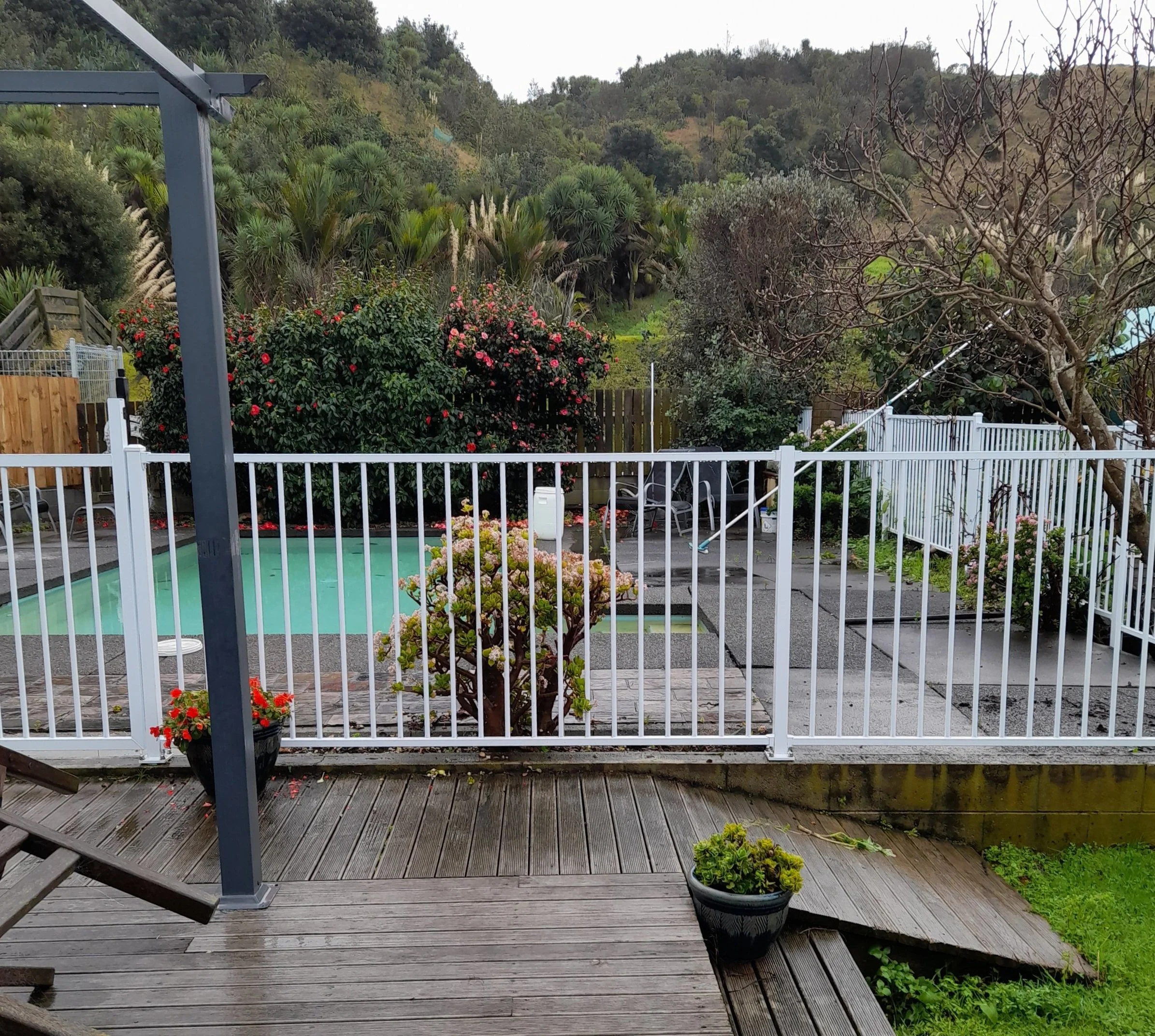 A backyard deck with a white fence, potted plants, a round pool, and a garden with flowers and trees, surrounded by hills and greenery.