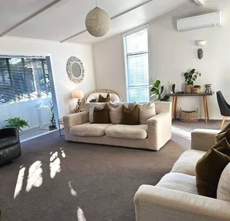 Living room with white sofas and brown pillows, a rug, a side table with a lamp, and a glass door leading outside. Walls are decorated with a mirror and a clock, with potted plants and a wooden sideboard.