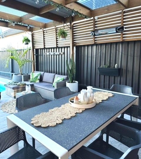 Outdoor patio with black table, six black chairs, gray sofa with green pillows, wooden coffee table, hanging potted plant, and decorative items on table and wall.