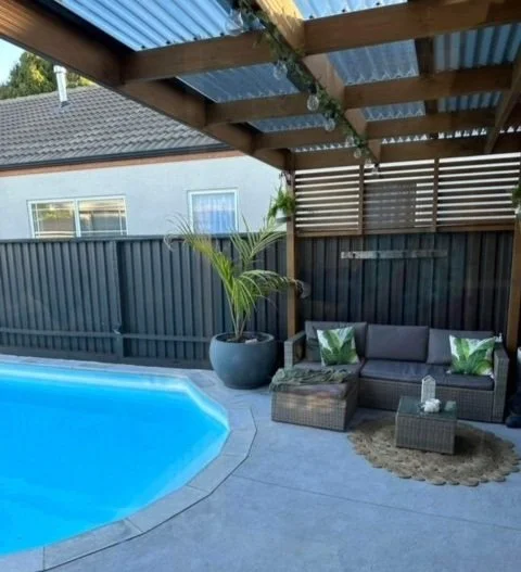 Backyard patio featuring a swimming pool, a seating area with a sofa, two green and white accent pillows, a small coffee table, and a large potted palm tree under a wooden pergola.