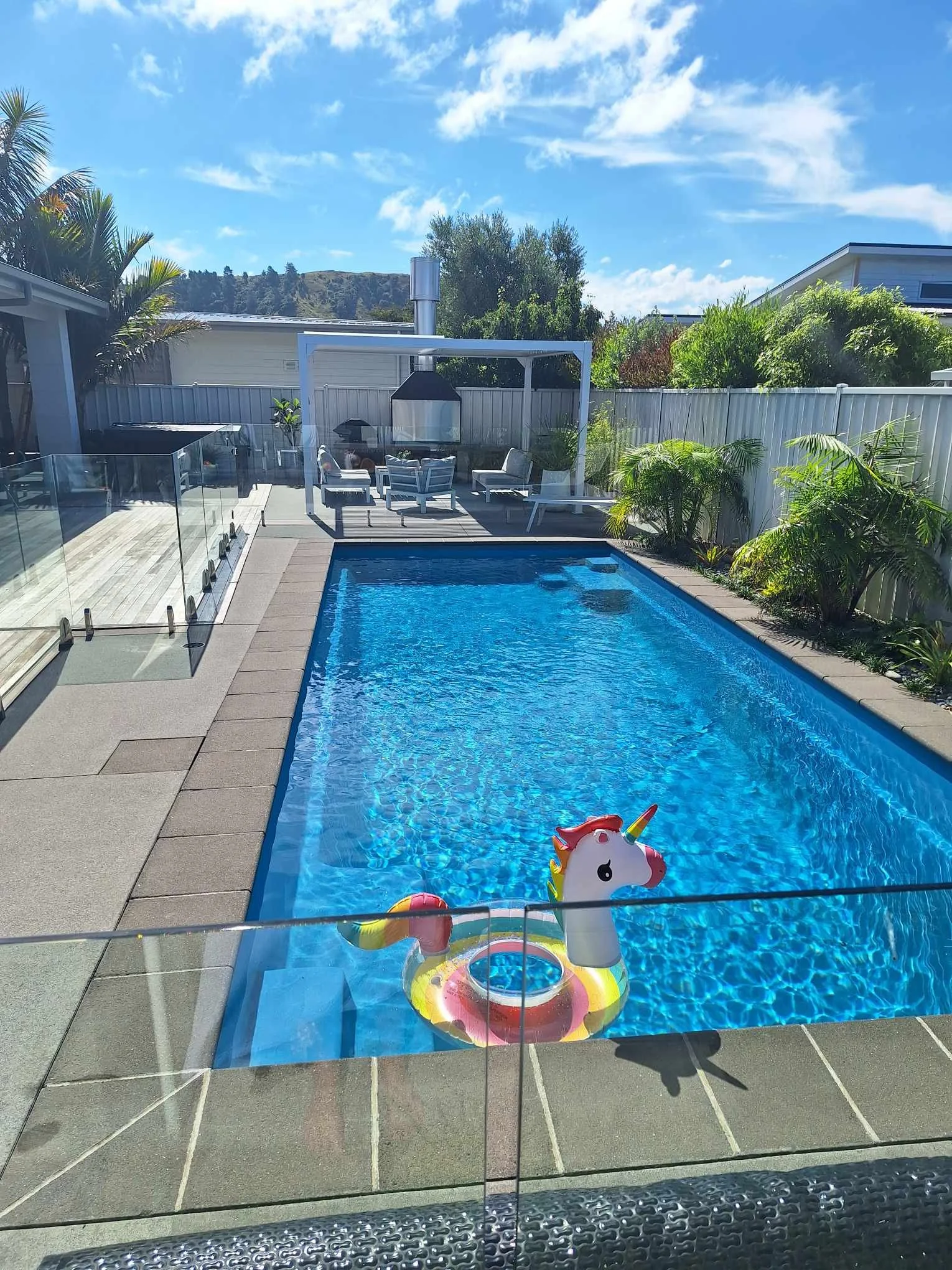 A backyard swimming pool with a colorful unicorn-shaped inflatable float, surrounded by glass fencing, lounge chairs, and lush greenery, under a sunny blue sky with some clouds.