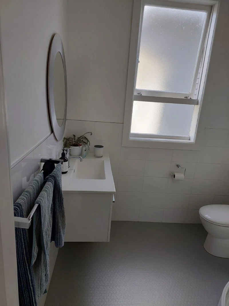 A small, minimalist bathroom with a white vanity, oval mirror, and a window letting in natural light.