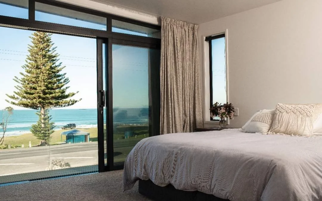 Bedroom with large window and sliding glass door overlooking a beach and ocean, with a pine tree outside, and a bed with white bedding and pillows.