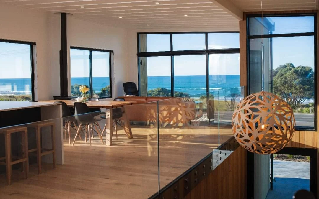 Interior view of a modern house with large windows overlooking the ocean, with a dining table, chairs, and a decorative hanging lamp.