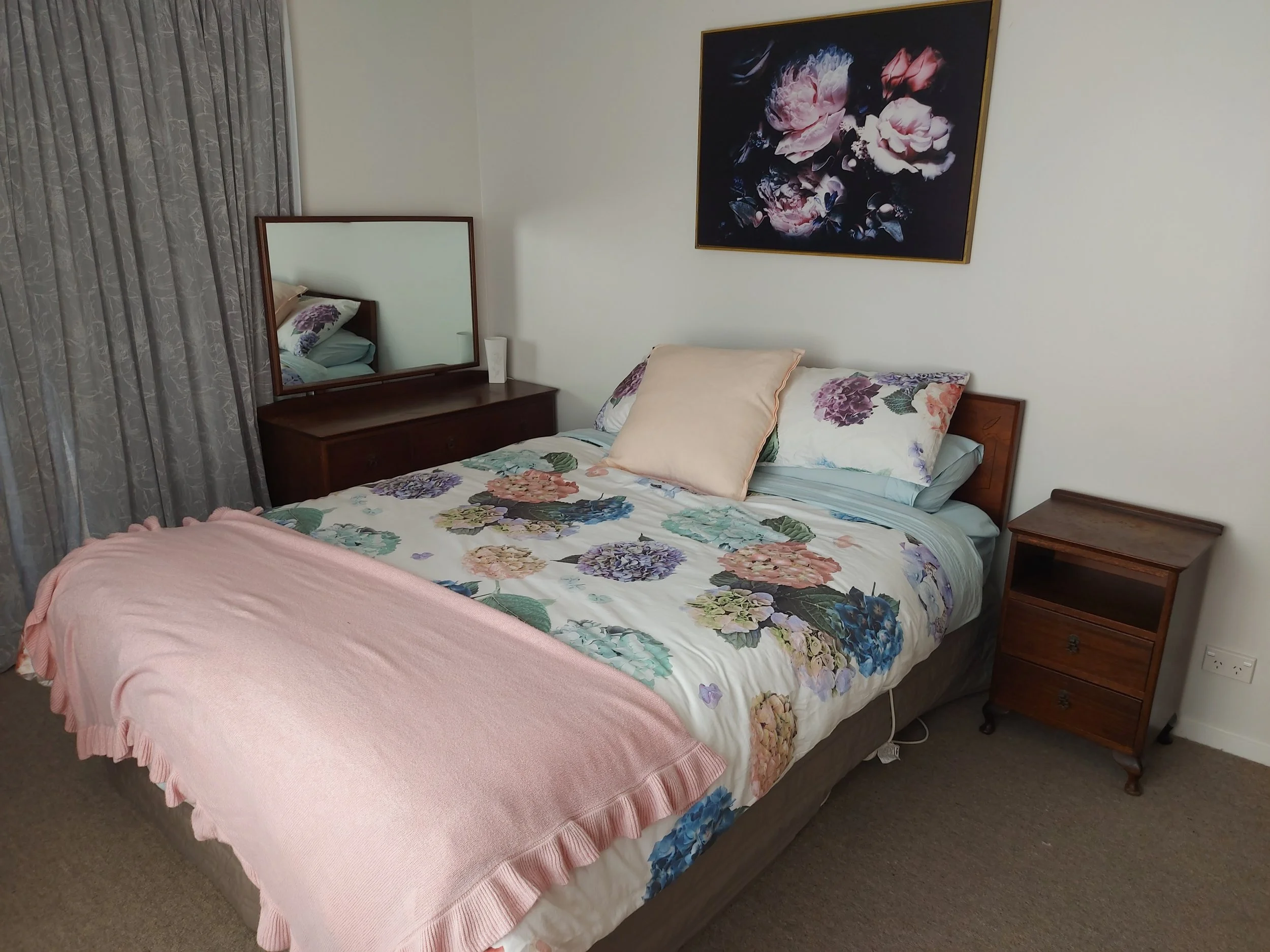 Bedroom with floral bedspread, pillows, a mirror on a wooden dresser, a floral painting on the wall, a nightstand, and gray patterned curtains