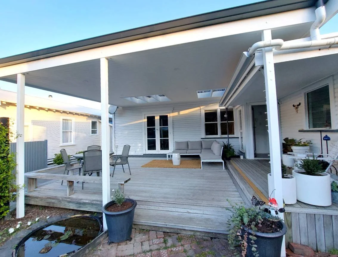 Backyard patio area with a wooden deck, outdoor seating including a sectional sofa and a dining table with chairs, potted plants, decorative items, and a small pond.