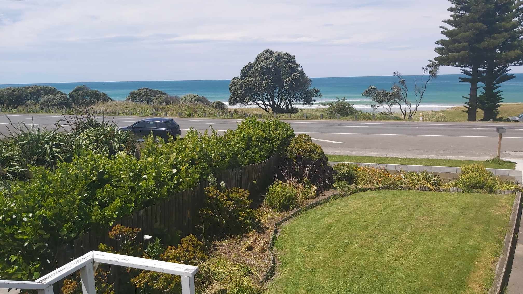 View of a coastal landscape with a garden in the foreground, a road with cars passing, and a beach and ocean in the background under a partly cloudy sky.