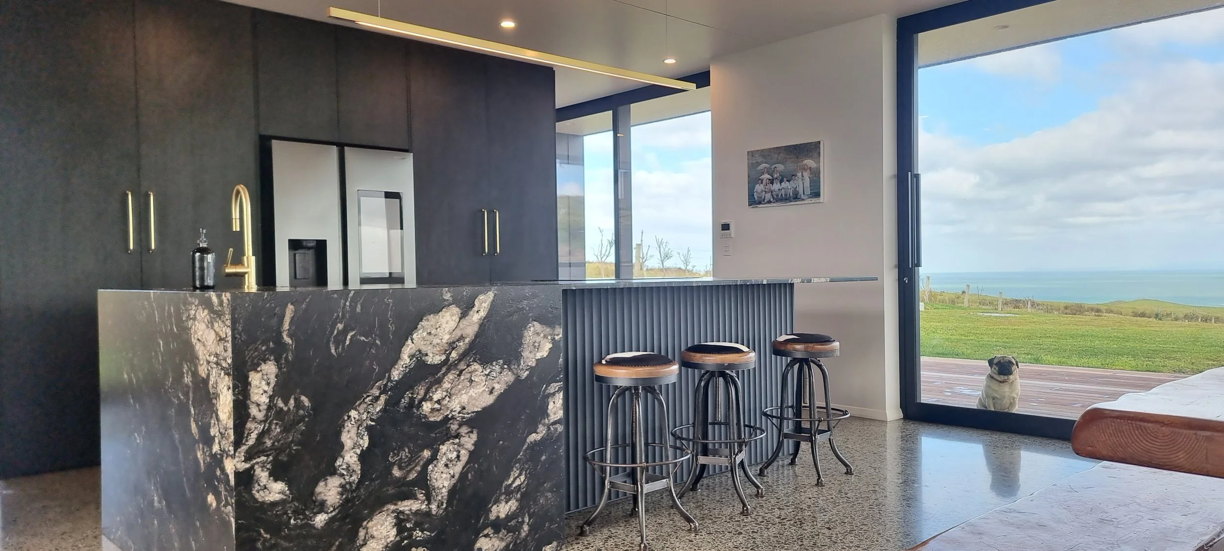 Modern kitchen with black cabinets, a marble island, four bar stools, large sliding glass door leading to a scenic outdoor view, and a dog sitting outside.