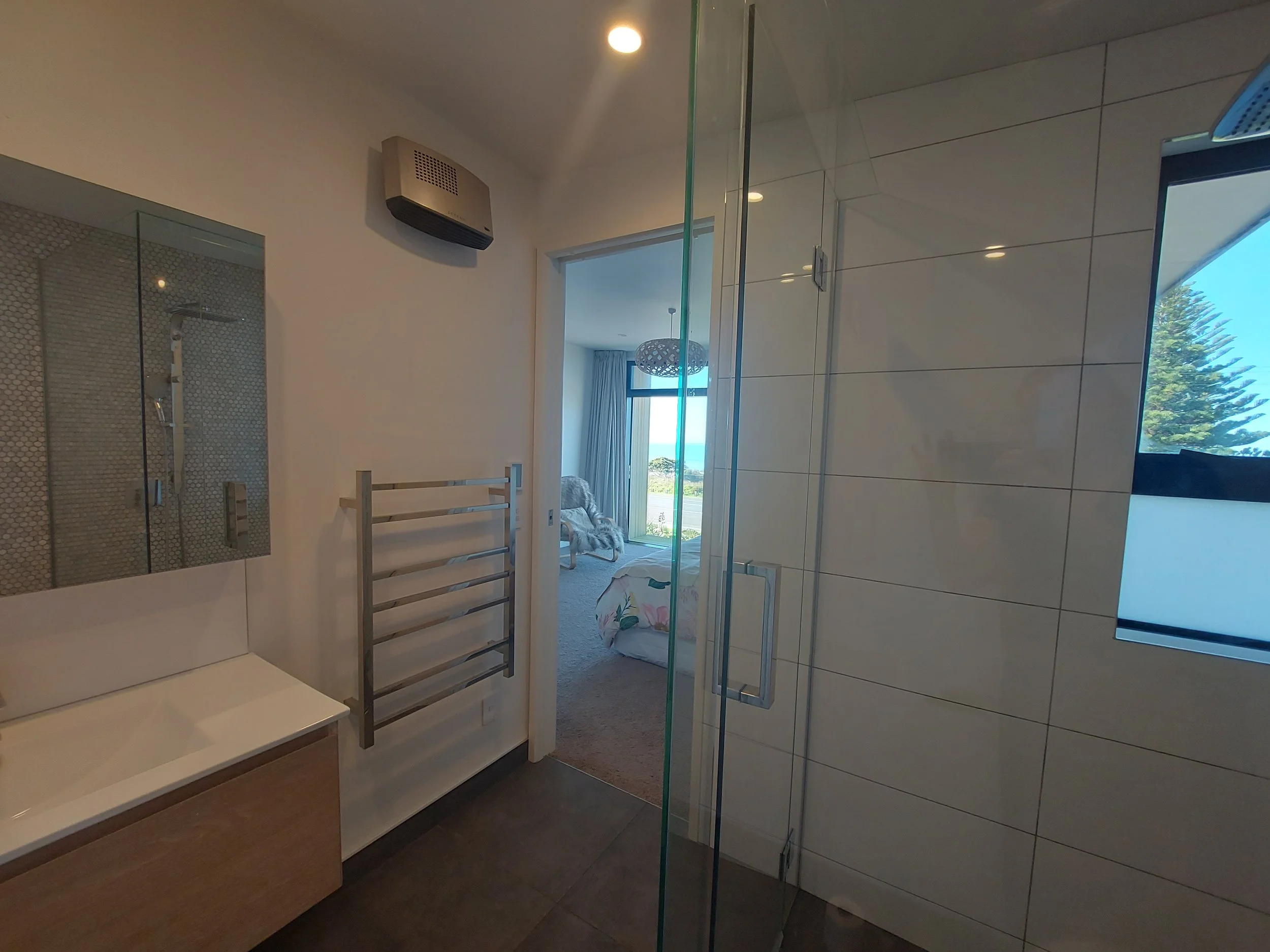 Bathroom with a glass shower, a wooden ladder towel rack, a window with a view of trees outside, and a mirror reflecting a room with a curtain and a decorative ceiling light.