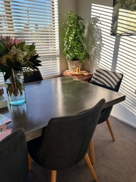 Dining room with a wooden table, black chairs, a vase of flowers, a tall potted plant, and blinds casting shadows on the wall.