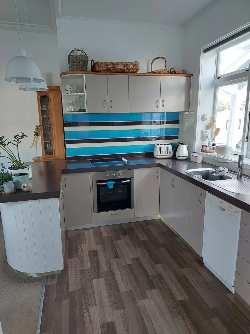 A kitchen with a black countertop, white cabinets, and a colorful striped backsplash. There are upper cabinets, a window above the sink, a toaster, a kettle, and baskets on top of the cabinets.