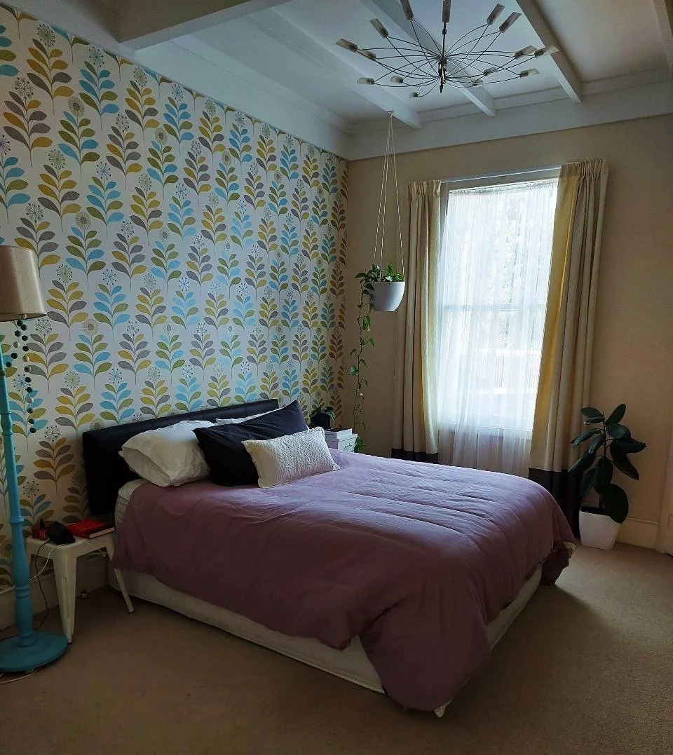 A bedroom with a bed that has a mauve comforter, black and white pillows, and a small decorative pillow. The wall behind the bed features a patterned wallpaper with blue, green, and beige leaves. is a white side table with items, potted plant hanging