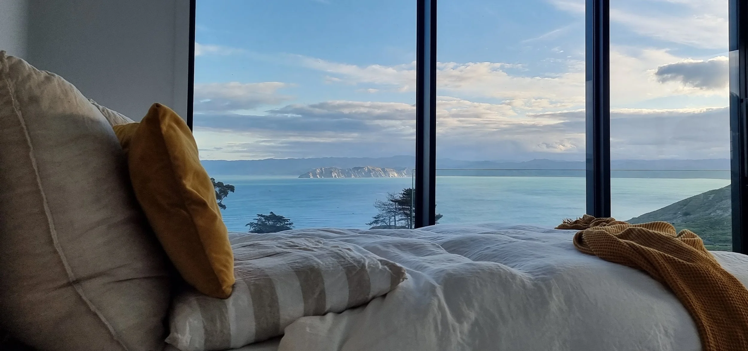 A cozy bed with beige pillows and a blanket, overlooking a scenic view of water, mountains, and a partly cloudy sky through large floor-to-ceiling windows.