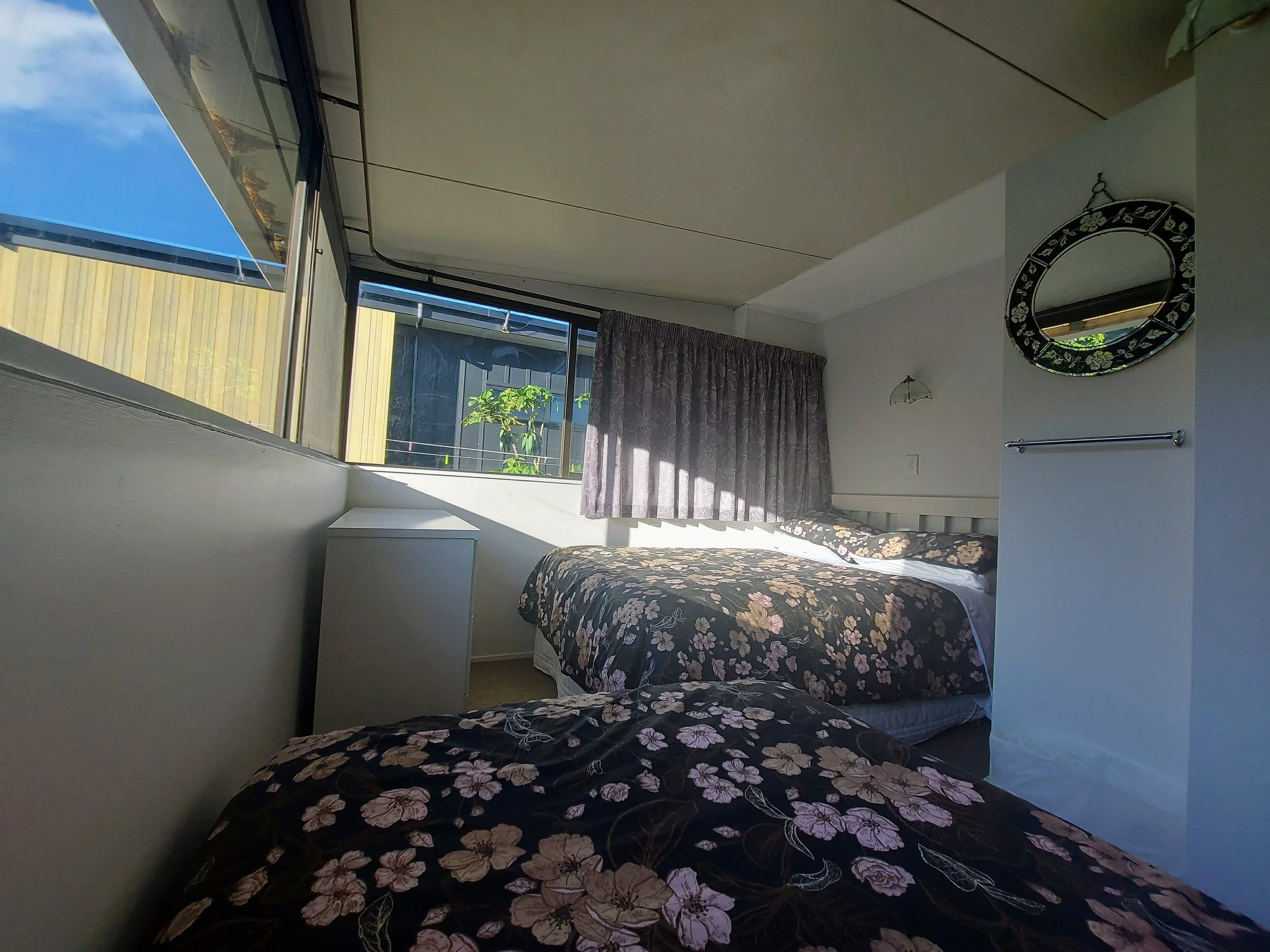 Interior of a bedroom with a floral bedspread, a white bedside table, a large window with curtains, a mirror decorated with flowers, and a balcony with plants outside visible through the window.