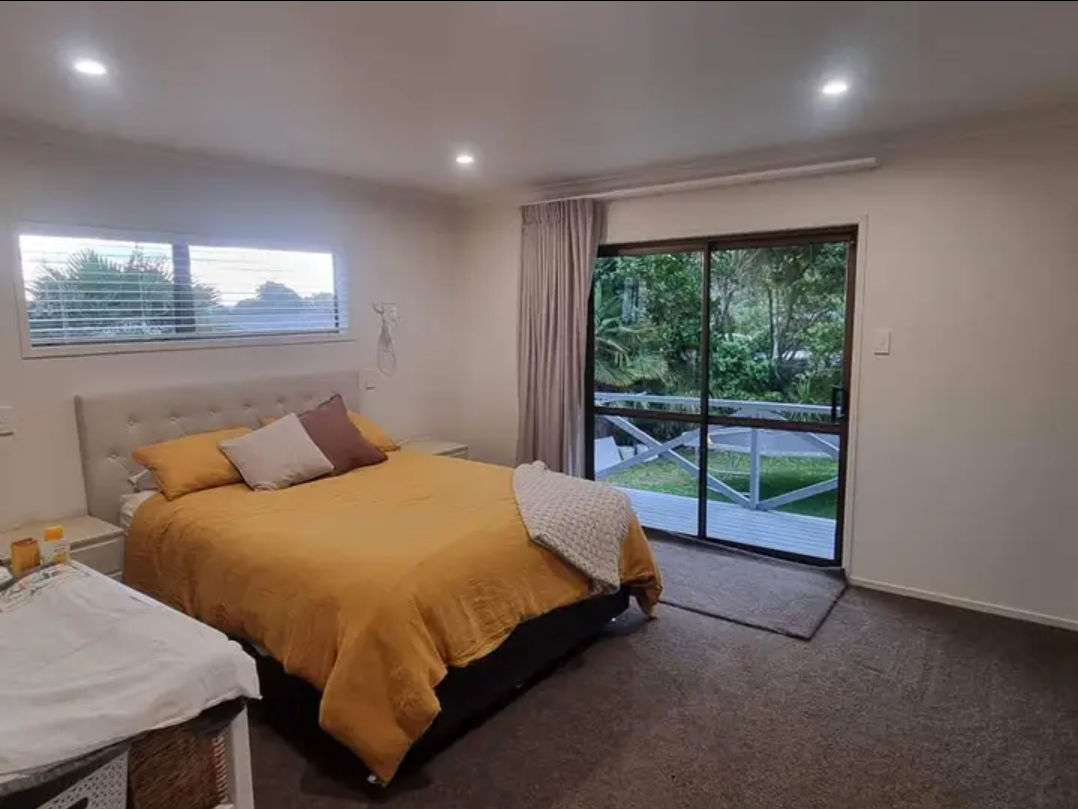 Bedroom with a bed covered in a yellow comforter, white and purple pillows, nightstand, sliding glass door leading to a outdoor balcony with greenery and trees outside.