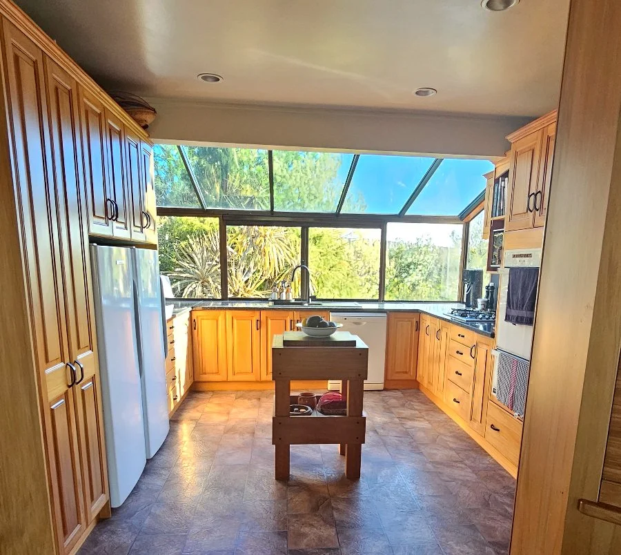 A bright kitchen with wooden cabinets, a small island, and large windows showing trees outside.