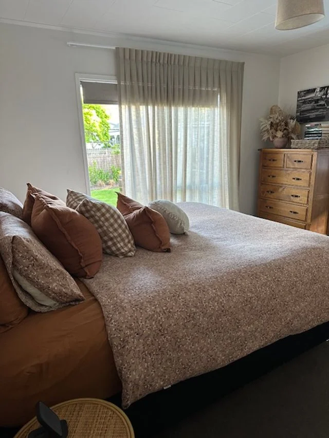 A bedroom with a bed covered by a quilt, several pillows, a window with curtains, a wooden dresser with decorative items and books, and a small circular side table with a lamp.