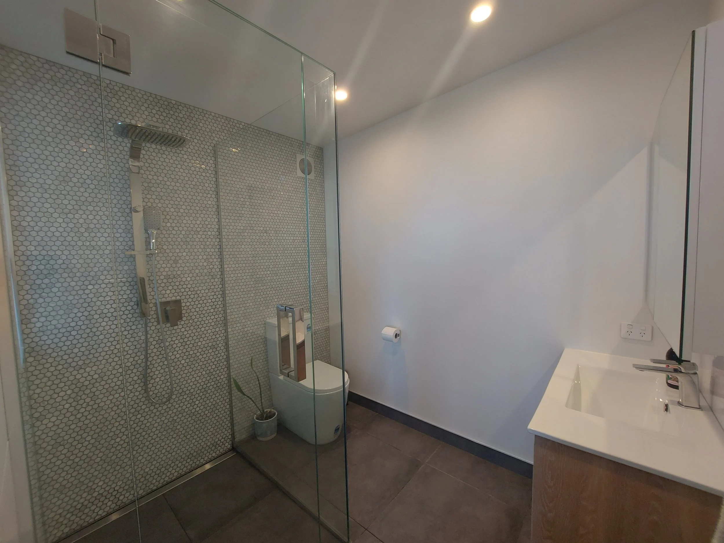 A modern bathroom featuring a walk-in shower with glass enclosure, hexagonal tile walls, and a white sink with a chrome faucet, illuminated by ceiling lights.