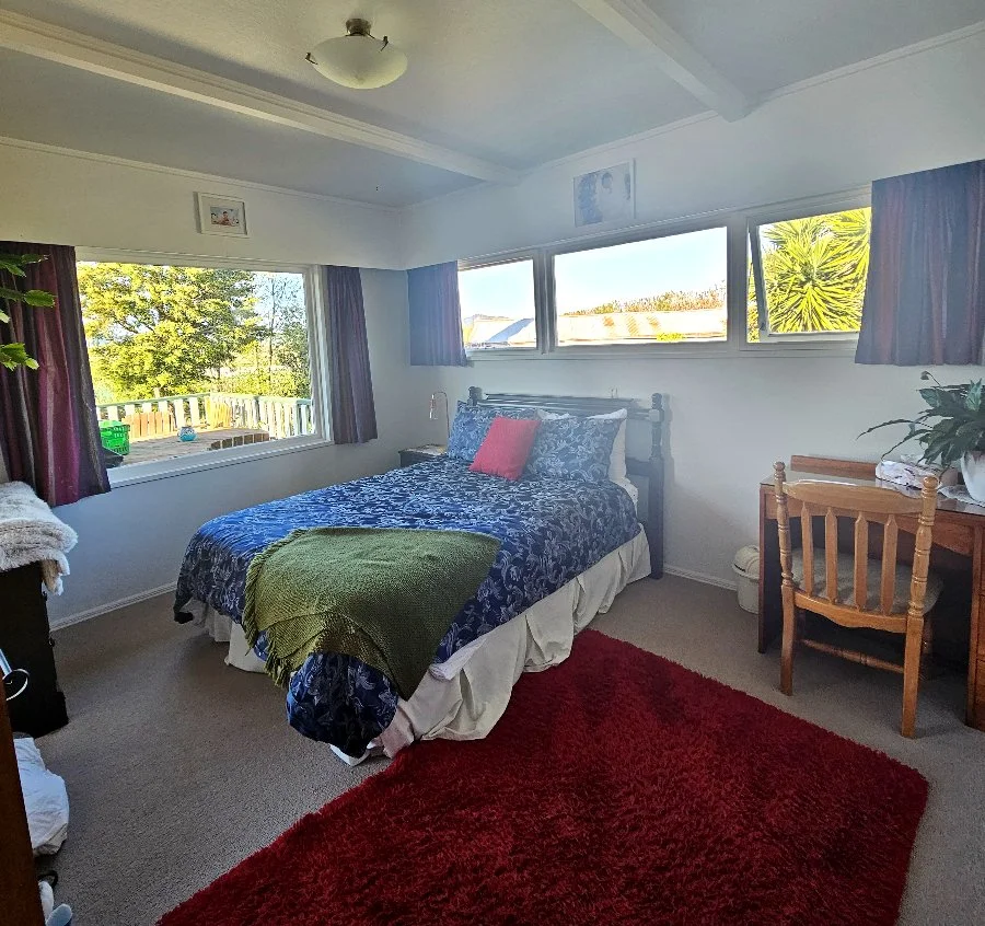 Bedroom with bed, colorful bedding, red rug, small desk, and view of trees outside the window.