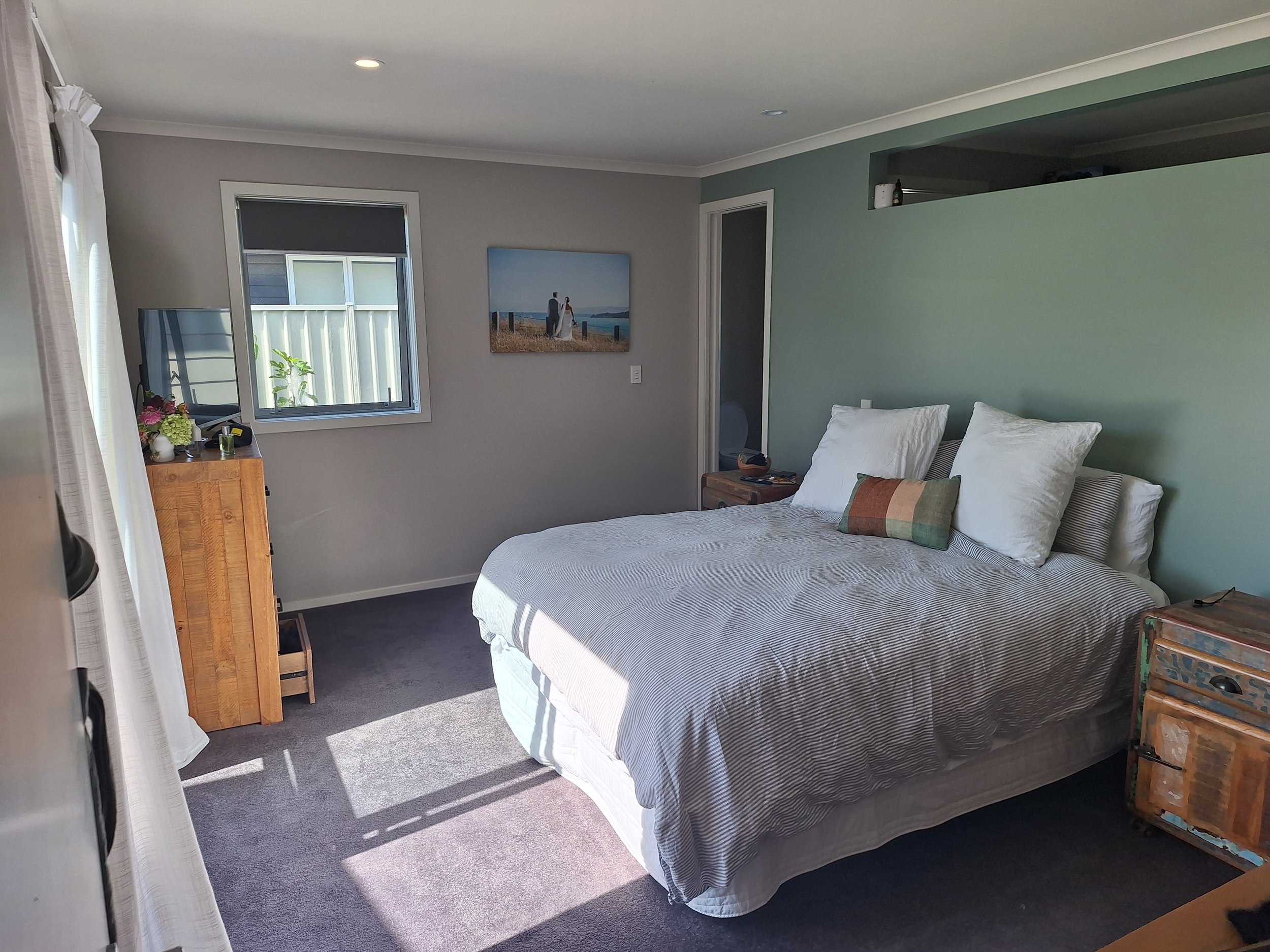 A cozy bedroom with a bed covered in a striped gray and white comforter and multiple pillows. There is a window with a black blind, a wooden dresser with flowers, a small painting of a couple on a beach, and a bedside table with a basket and various 