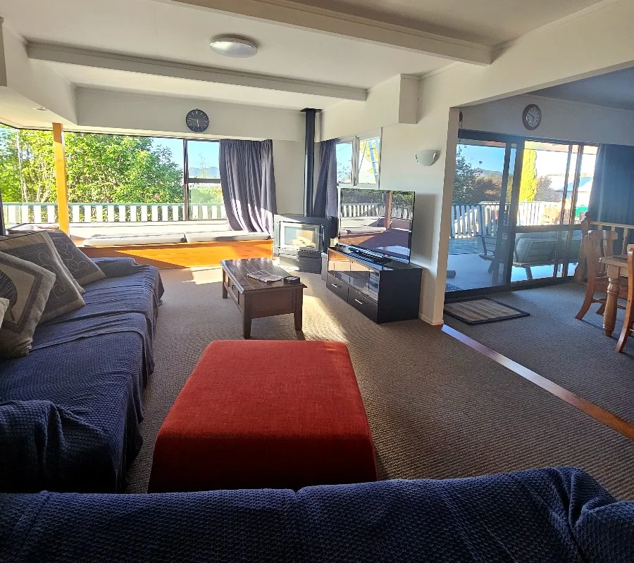 Living room with couches, a TV, a wood stove, and a large window overlooking greenery and a deck.