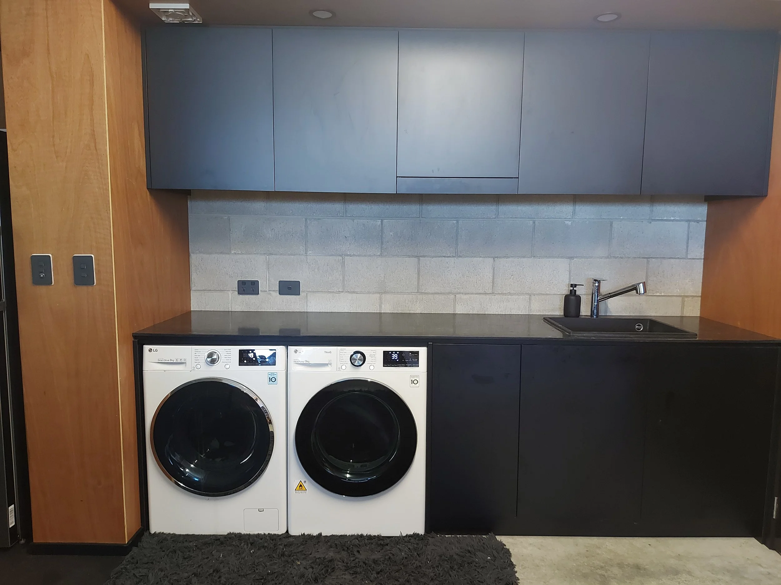 A laundry room with a wooden wall on the left, blue upper cabinets, gray concrete block wall, black countertops, a black sink with a silver faucet, and two front-loading white washing machines with digital displays and black doors. There are black el