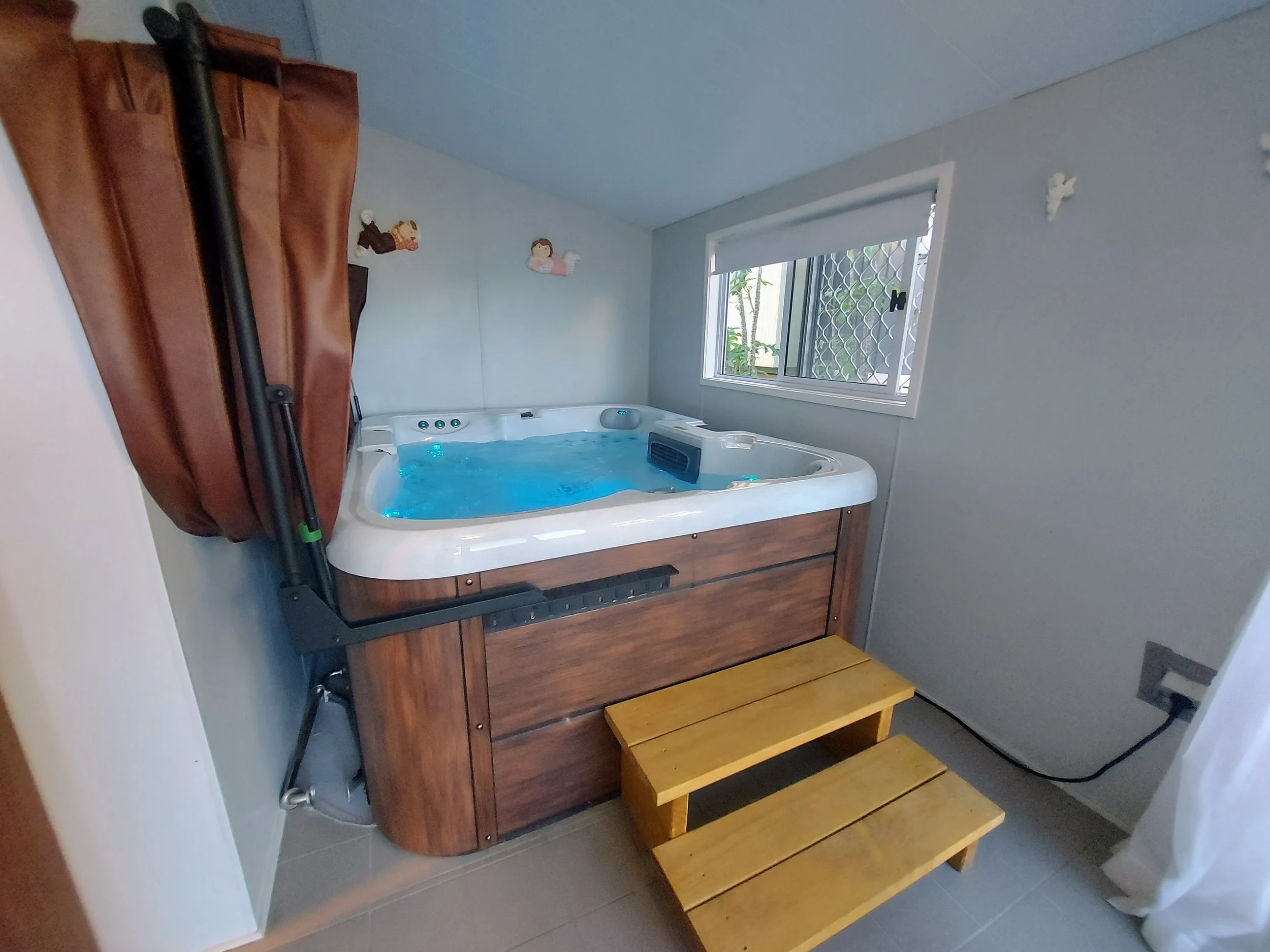 A corner of a room with a hot tub, small decorations of angels on the wall, a window with a mesh screen, a shower head, a brown curtain, and a wooden step stool.