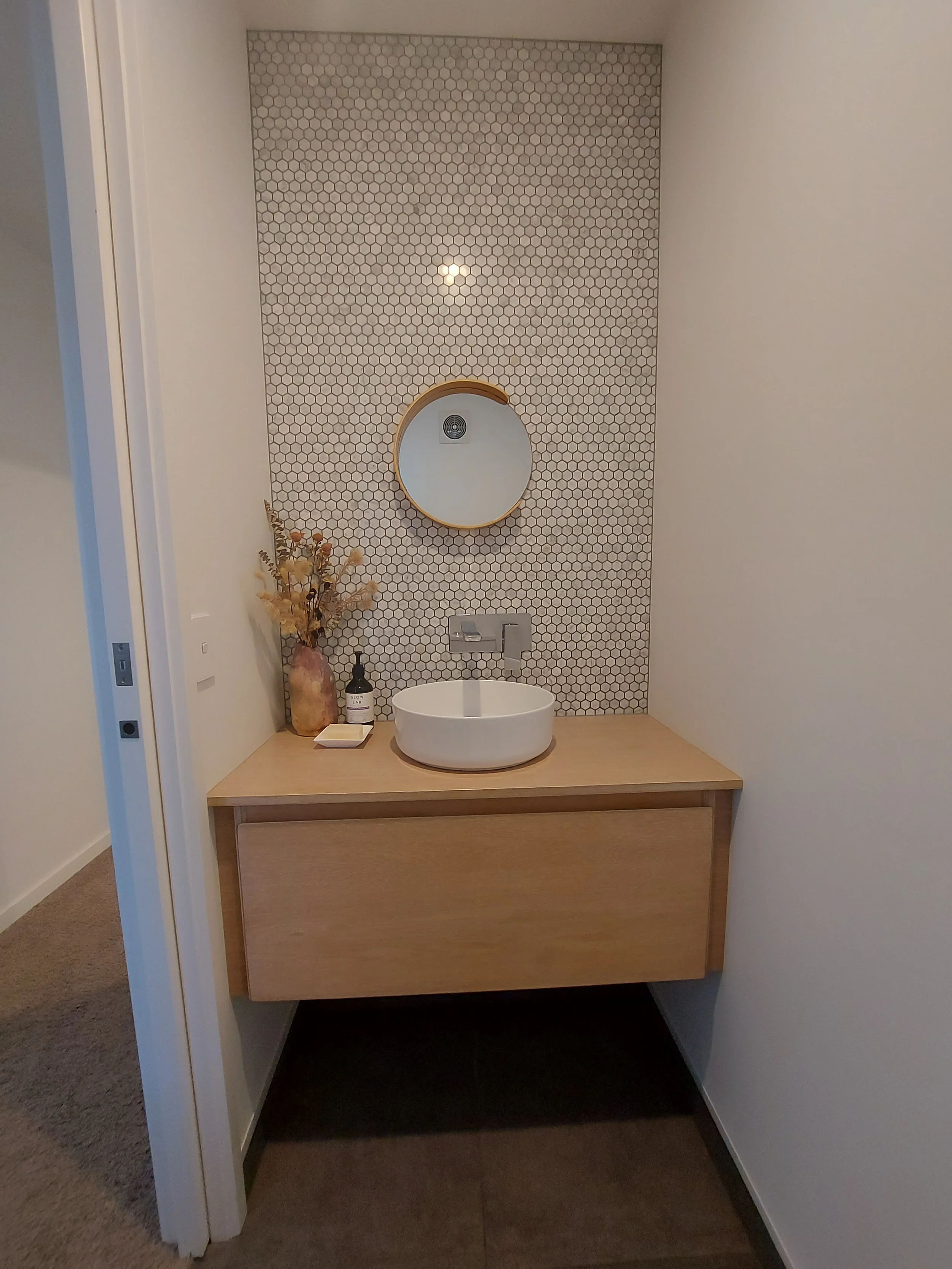 A modern bathroom vanity area featuring a white vessel sink on a wooden cabinet. There is a round mirror above with a wooden frame, and a hexagonal tile accent wall behind. Decor includes a dried flower arrangement in a large vase, a soap dispenser, 