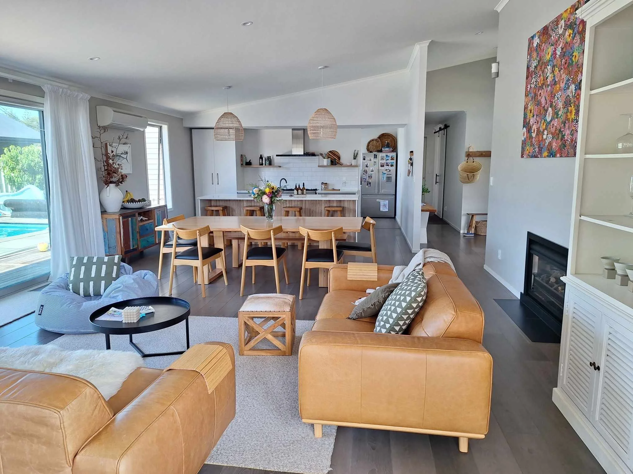 Open-concept living and kitchen area with wooden dining table, tan leather sofa, hanging wicker lamps, and exterior through sliding glass door.