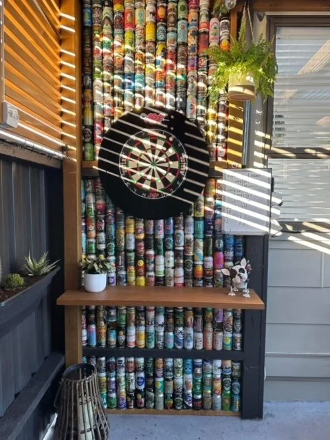 A dartboard with a colorful background of stacked cans, a small shelf with plants and decorative items, and sunlight casting shadows through nearby blinds.