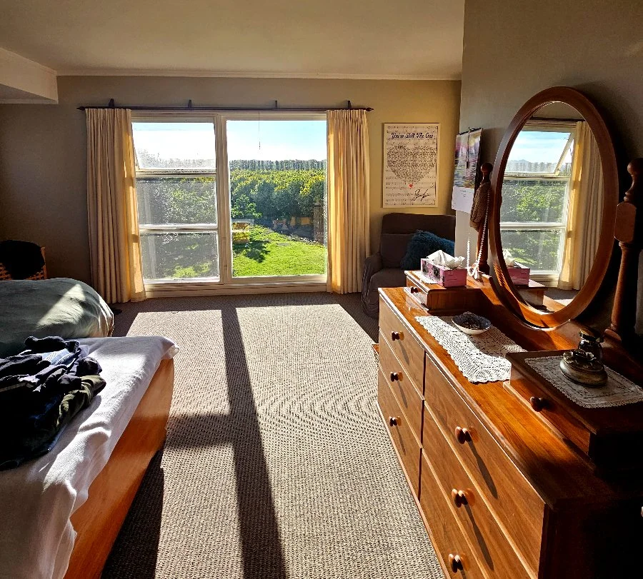 A bedroom with a large window showing a green landscape outside. The room has a bed, a wooden dresser with a large oval mirror, and a small armchair near the window. Light curtains frame the window, and sunlight illuminates the room.