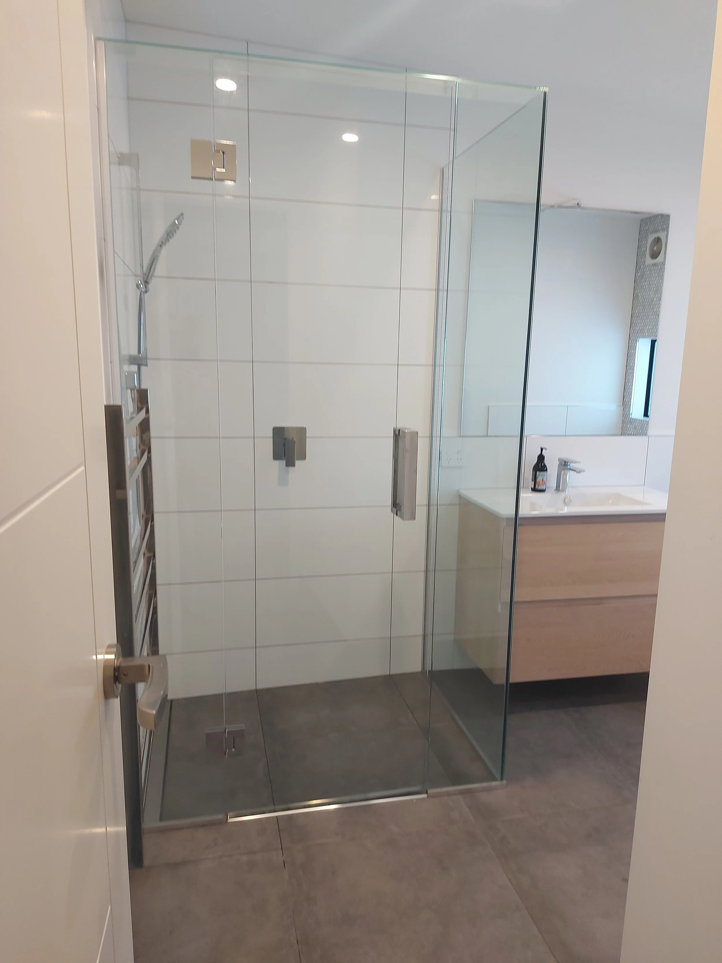 A modern bathroom with a glass shower enclosure, white tiles, a small mirror, a sink, and a soap dispenser.