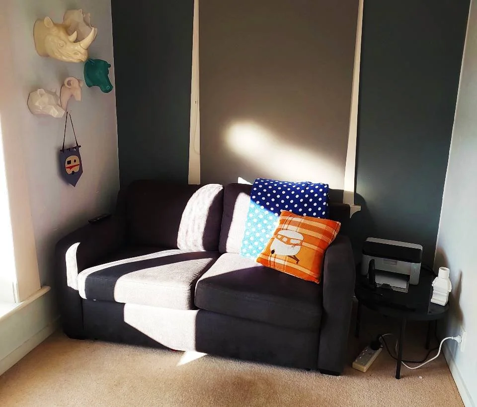 A dark gray sofa with three cushions: one with white polka dots on a blue background, one with an orange and white striped pattern with a camper van illustration, and a plain white cushion. To the right, a black round side table holds a printer and a