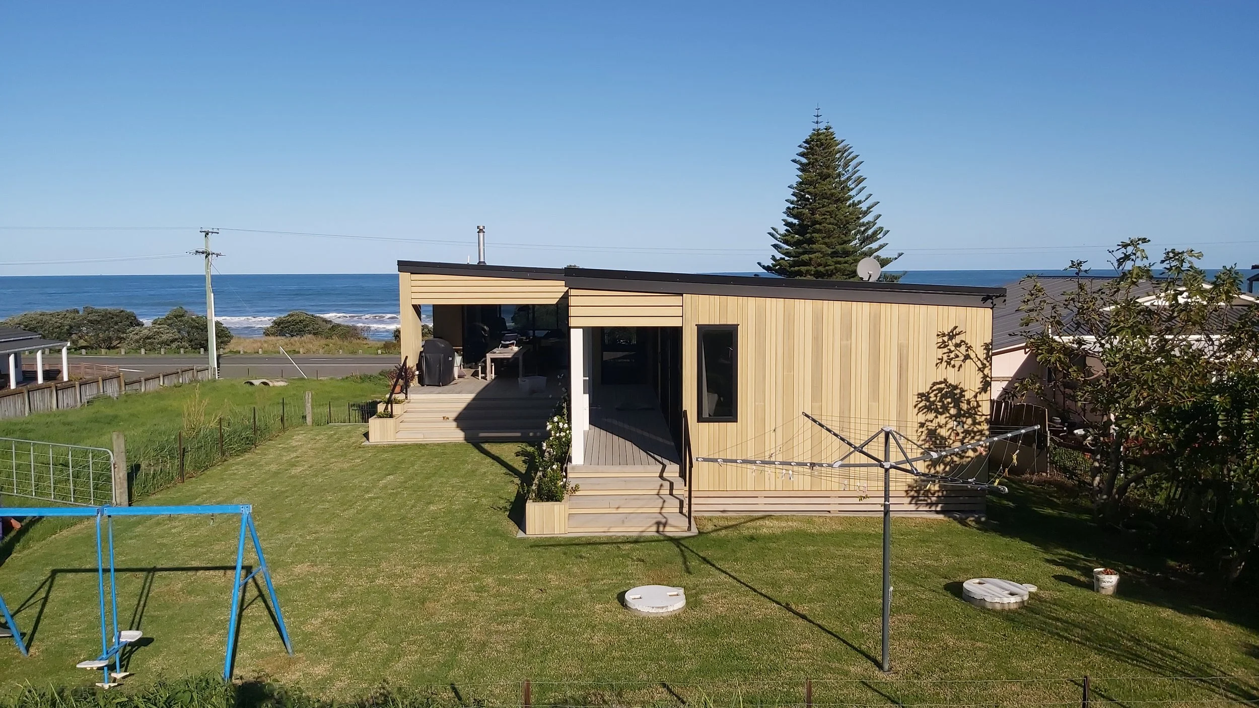 A modern wooden house by the ocean with a small lawn, a swing set, laundry drying lines, and a clear blue sky.