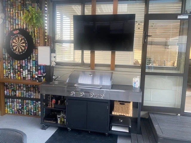 Indoor space with a dartboard on the left, a dartboard cabinet, a shelf filled with cans, a counter with a grill and a bottle of hand sanitizer, a flat-screen TV above, and a glass door on the right.