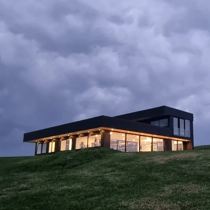 Modern house with large glass windows, situated on a grassy hill, illuminated from inside, under a cloudy sky.