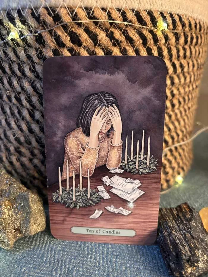 A woman sits at a table with her head on her hands, an expression of exhaustion and distress on her face while she is surrounded by receipts and bills. 10 candles surround her in a festive wreath, 5 on each side of her but none are lit