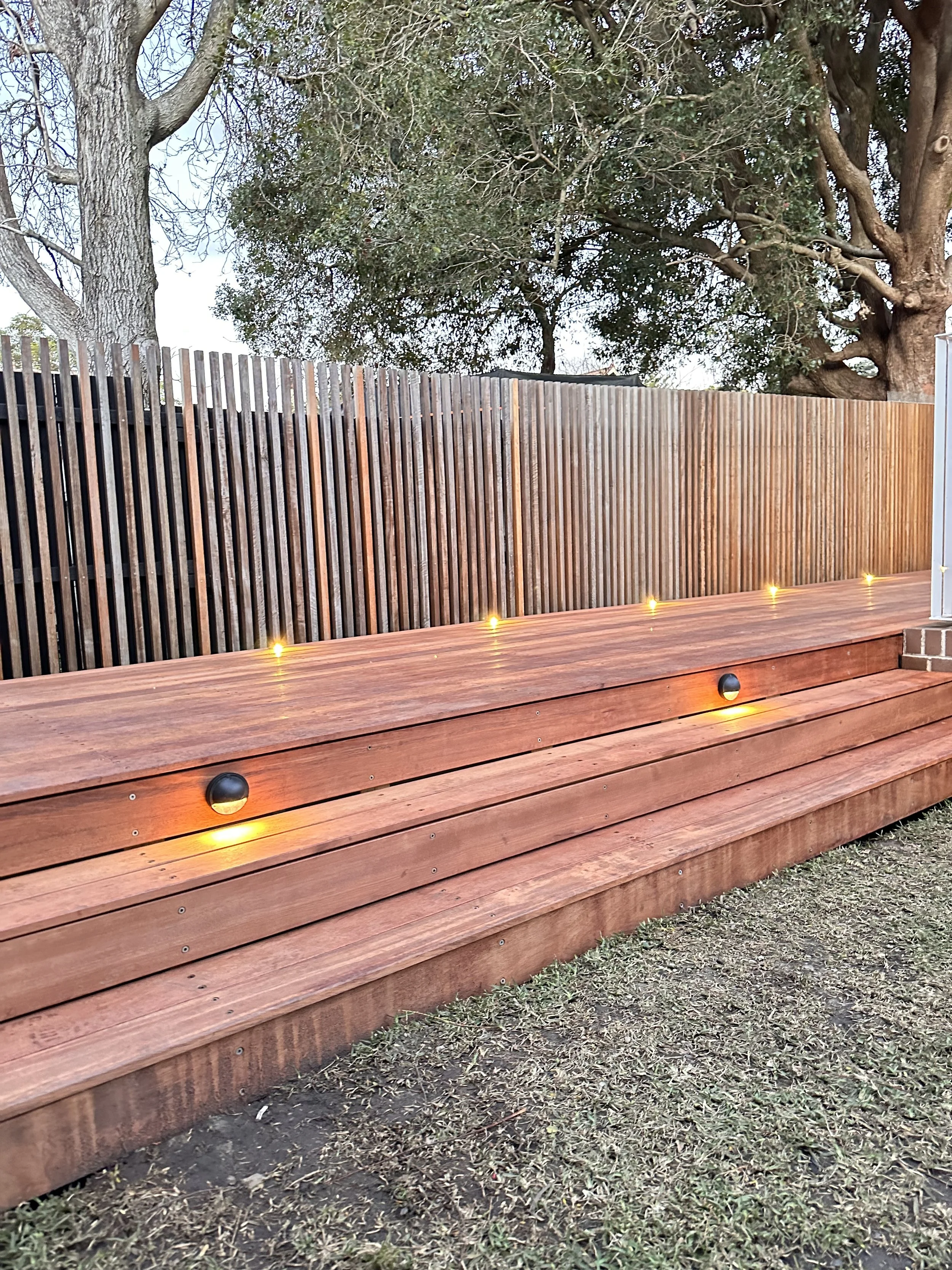 Wooden outdoor deck with built-in lighting, surrounded by a wooden fence and trees in the background.