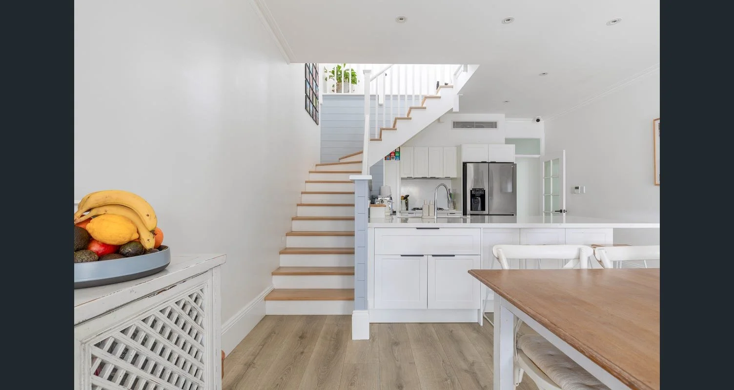Modern kitchen with white cabinets, stainless steel refrigerator, and wooden staircase leading to an upstairs area.