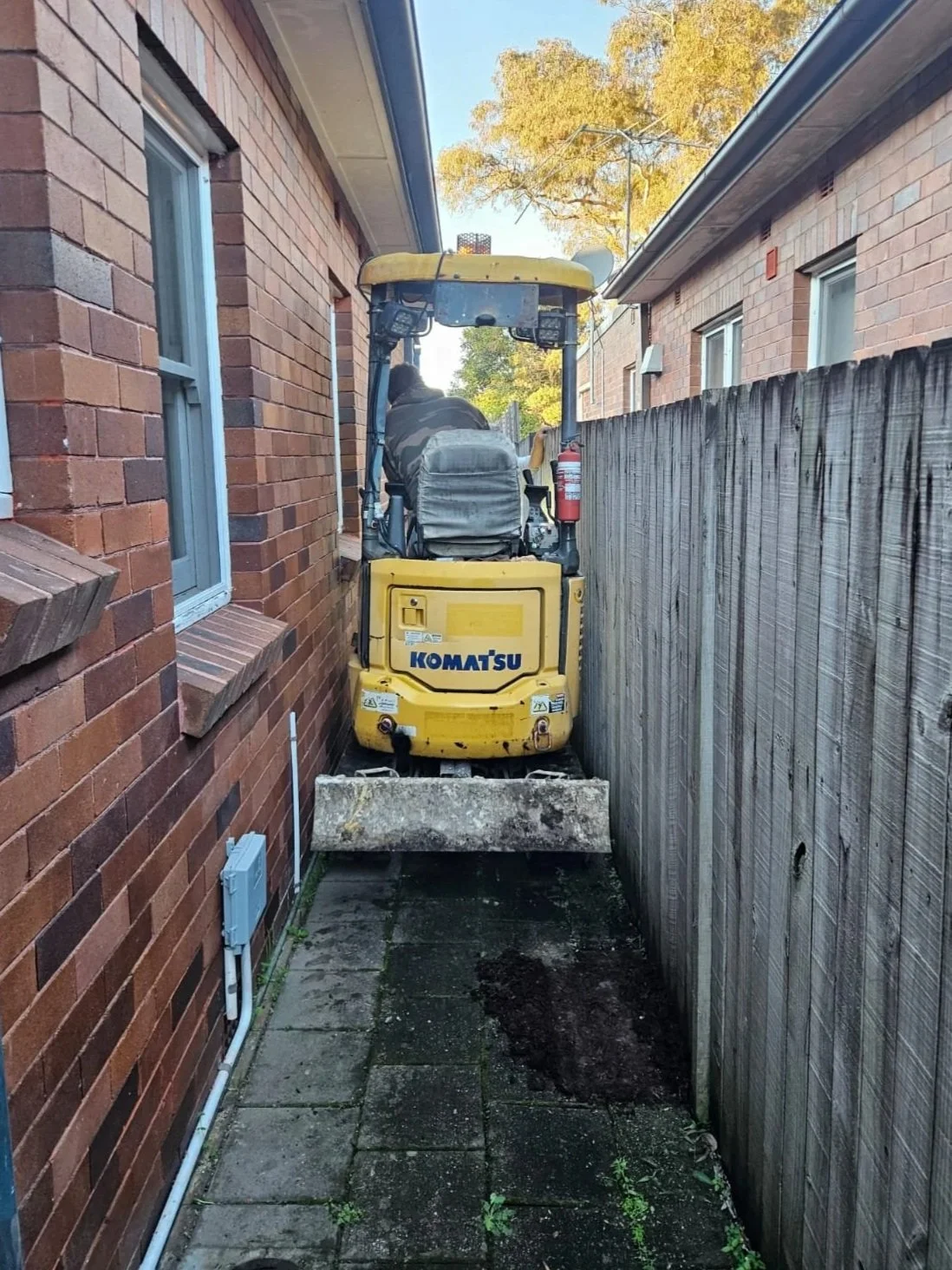 A person operating a yellow Komatsu mini excavator in a narrow alley between two brick houses, with a wooden fence on one side and a brick wall on the other, during daylight.