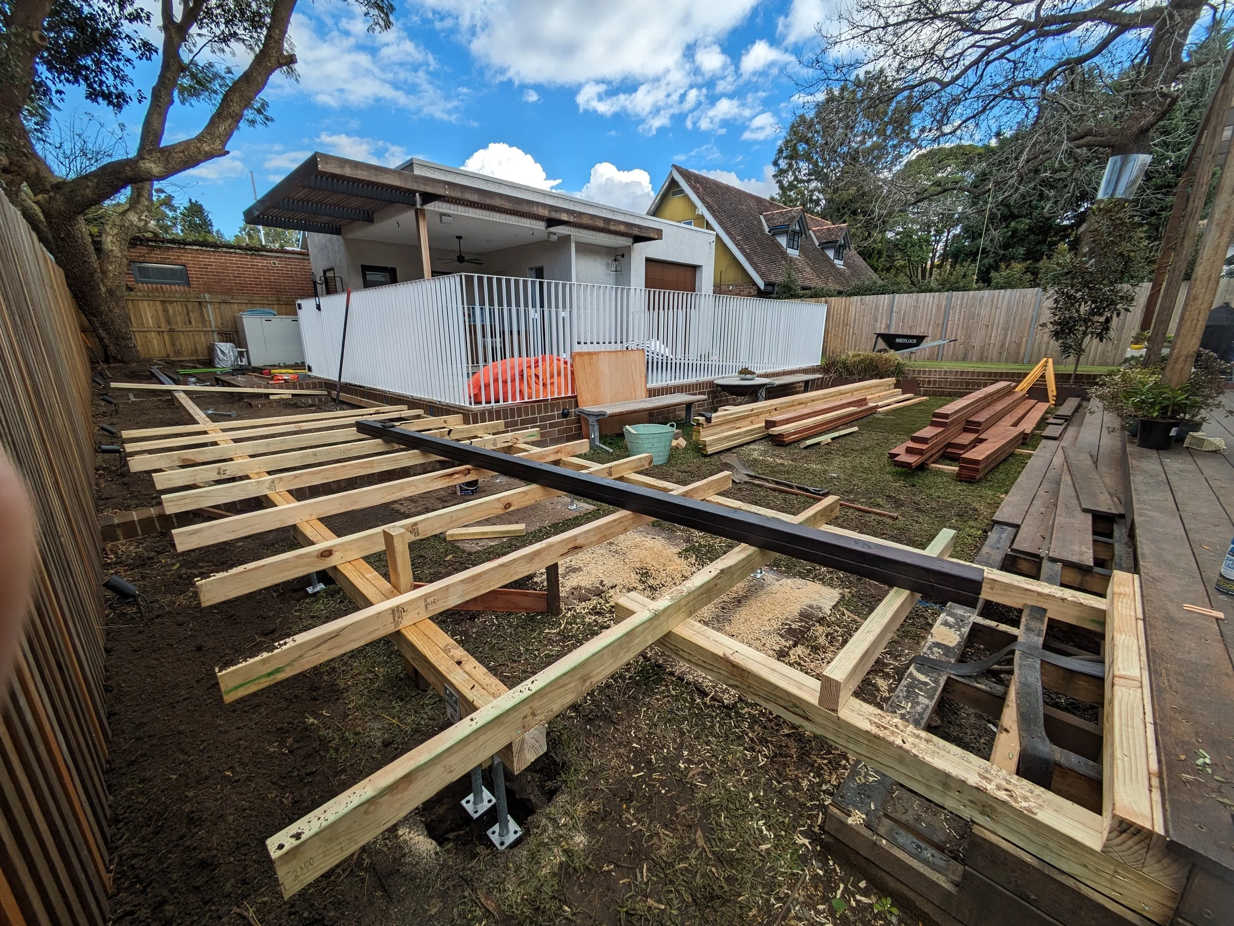 Backyard under construction with wooden framing for a deck or patio, surrounded by a wooden fence, with a tree on the left, nearby houses, construction tools, and materials scattered around.