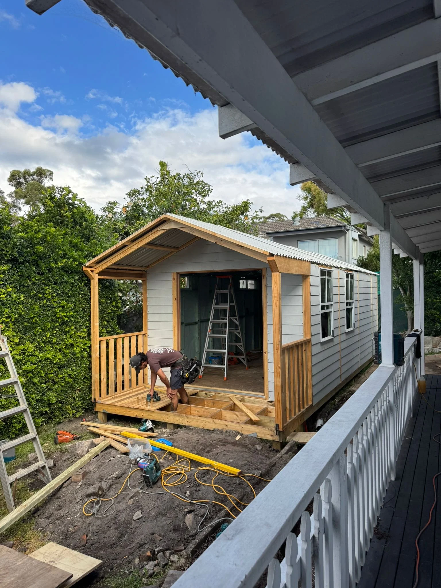 Person working on a small wooden porch in a backyard, using tools and with construction materials and tools around, during daytime.