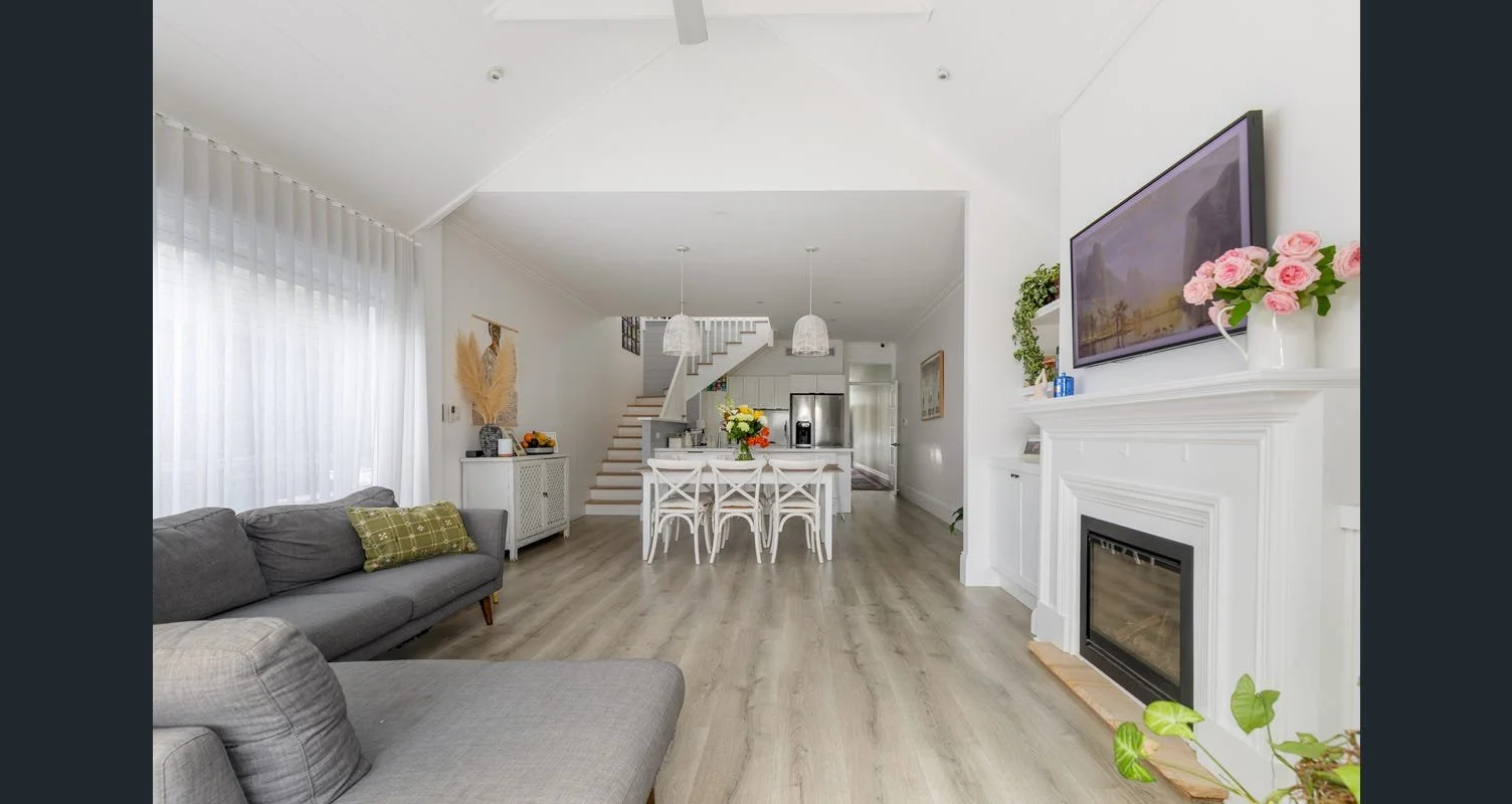 Bright living room with gray sofas, white walls and curtains, and a fireplace with a flat screen TV and pink roses. Dining table with white chairs, kitchen with stainless steel fridge, and staircase in background.