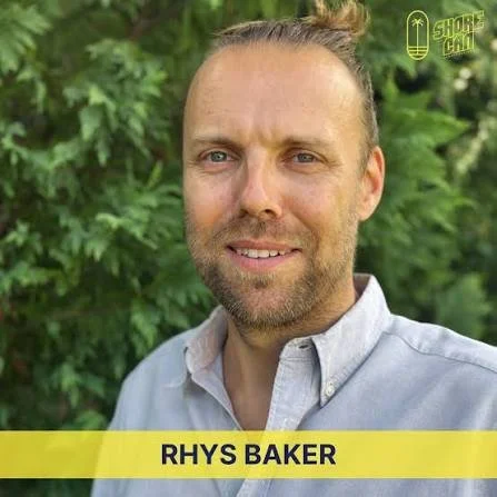 Man with light brown hair and a beard, wearing a light gray collared shirt, standing outdoors with green foliage in the background, smiling at the camera, with a yellow banner at the bottom displaying the name Rhys Baker, and a logo in the top right corner that says Shore Chil.