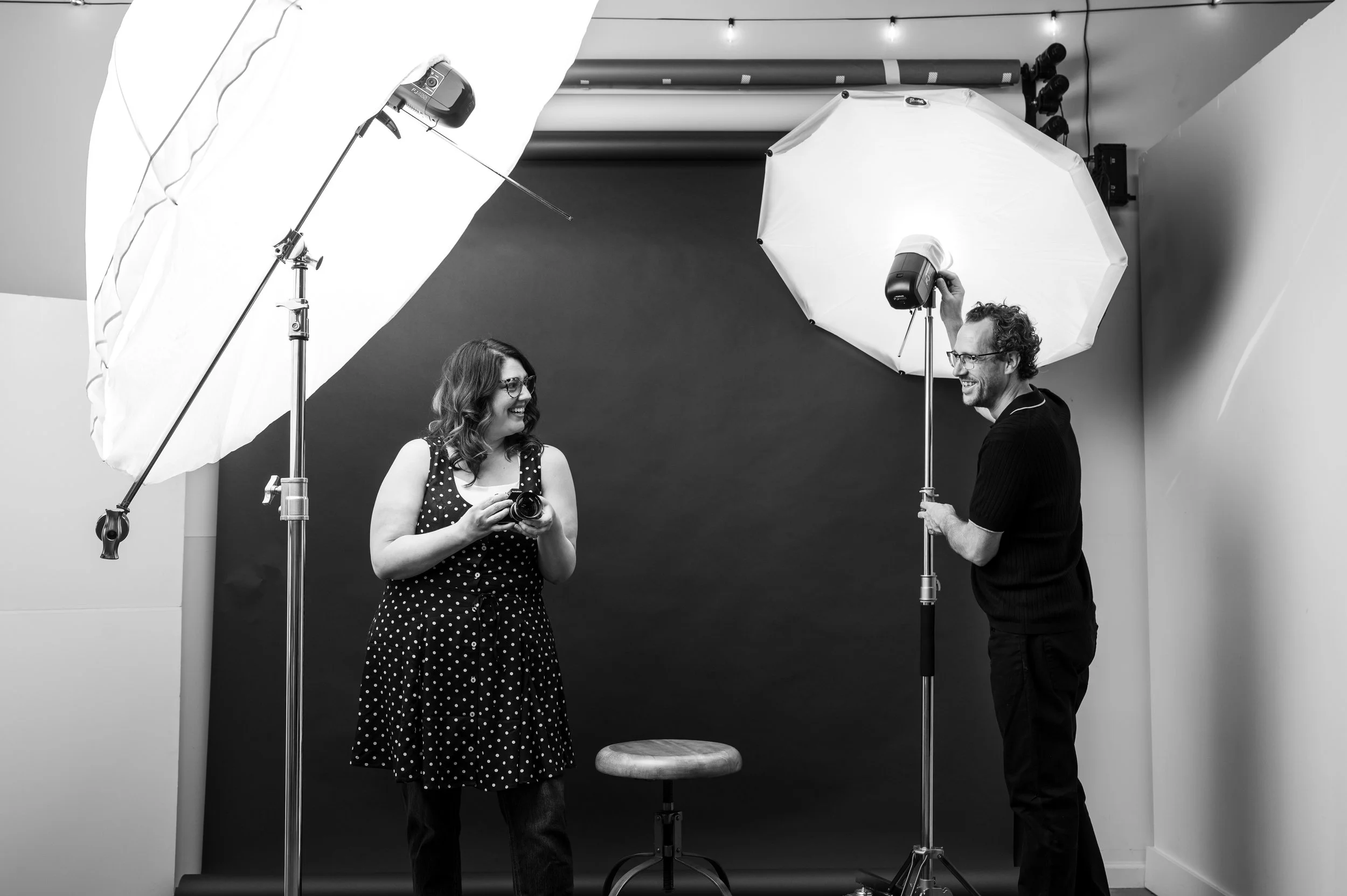 Black and white photo of a woman and a man in a photography studio. The woman is holding a camera and smiling, wearing glasses and a polka dot dress. The man is adjusting a large umbrella light, smiling. There are two large umbrellas and a backdrop behind them, with studio lights and equipment visible.