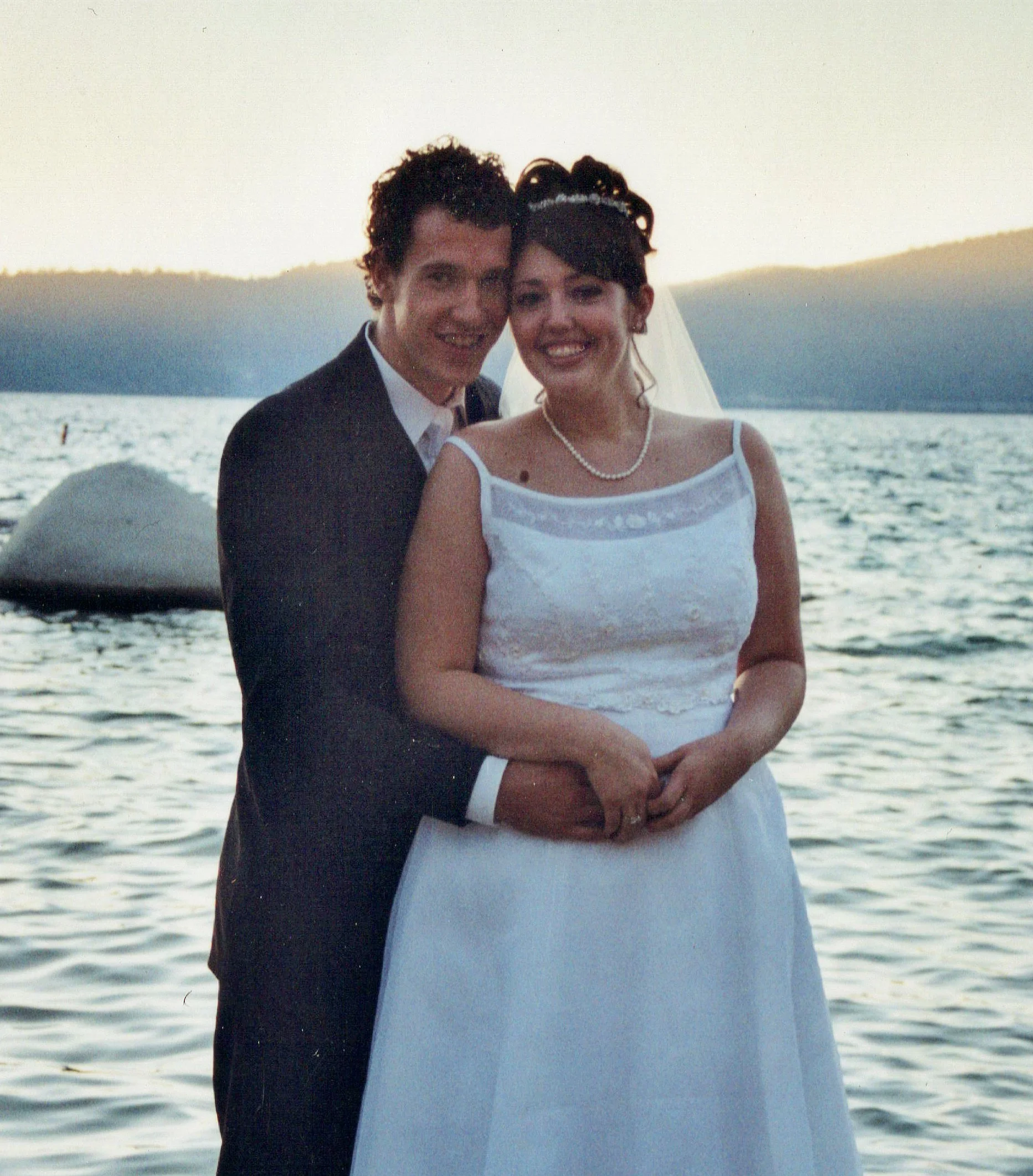 A smiling couple in a forest embraces, touching foreheads affectionately.
