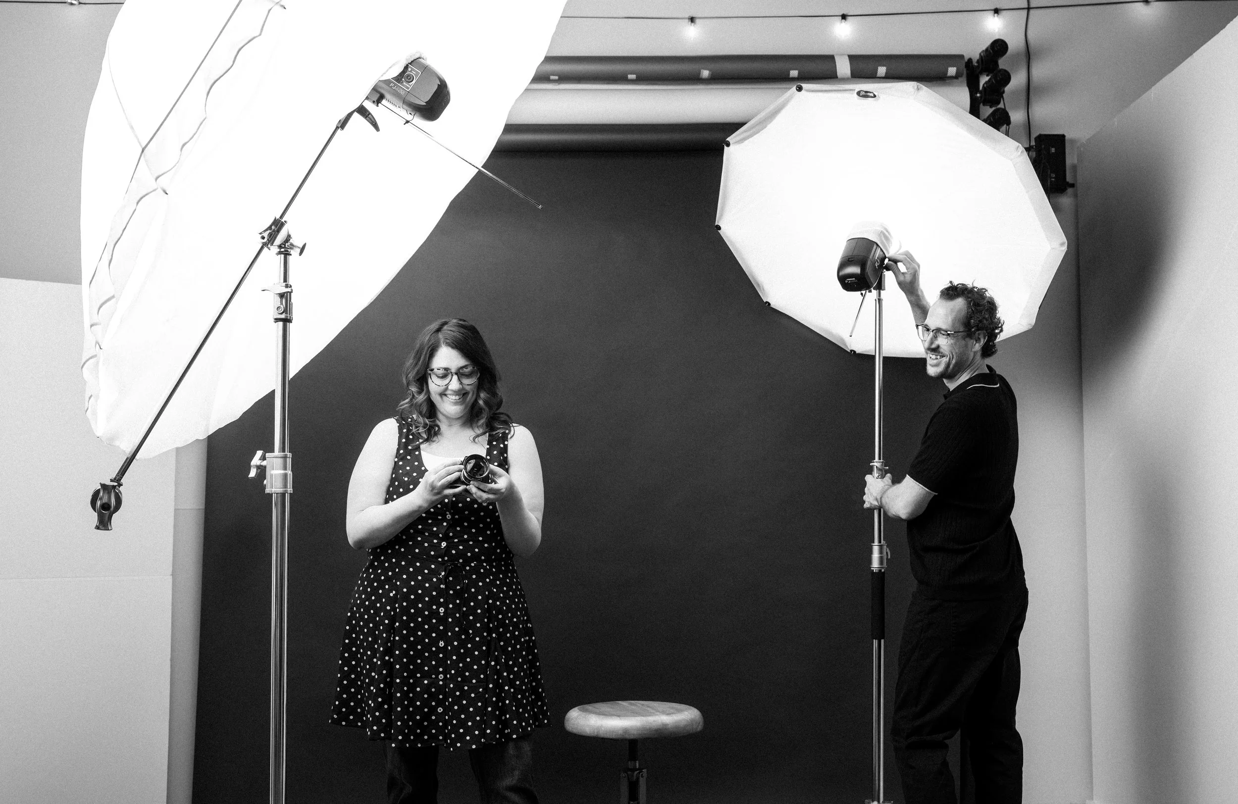 Two people standing in a photography studio. The woman on the left is looking at her camera and smiling, while the man on the right is adjusting a large umbrella light. There are two large studio lights with umbrellas set up, one on either side of the scene, and a small stool in the center.