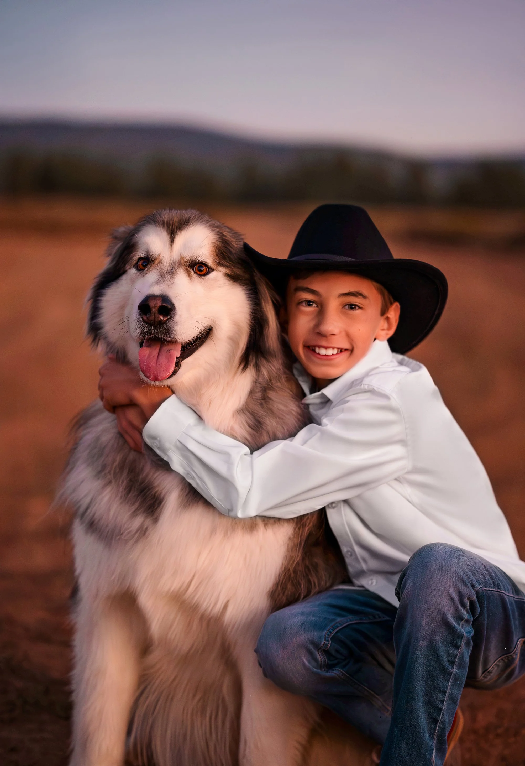 A young boy wearing a black cowboy hat and a white shirt hugging a large, fluffy, black-and-white Siberian Husky dog outdoors at sunset.