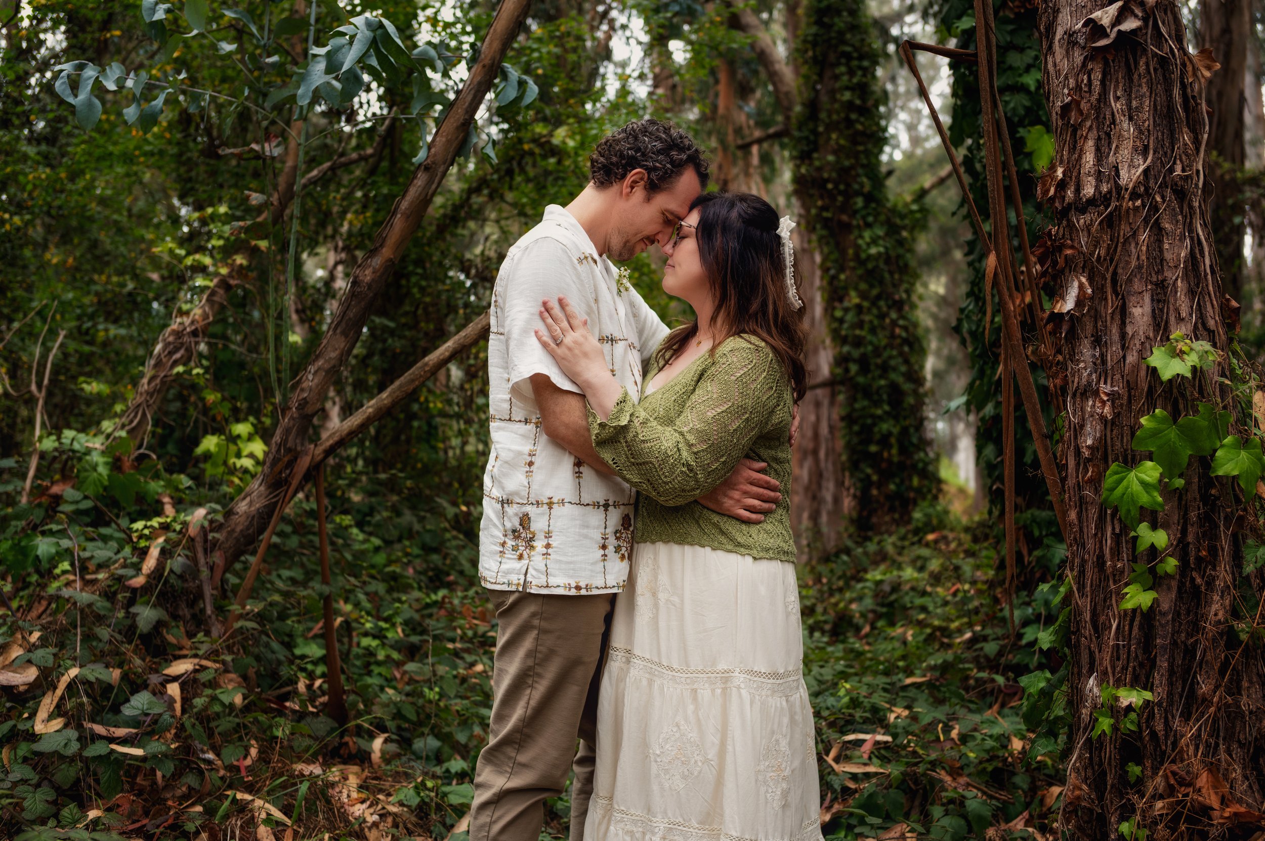 A smiling couple in a lush green forest embraces, touching foreheads affectionately.