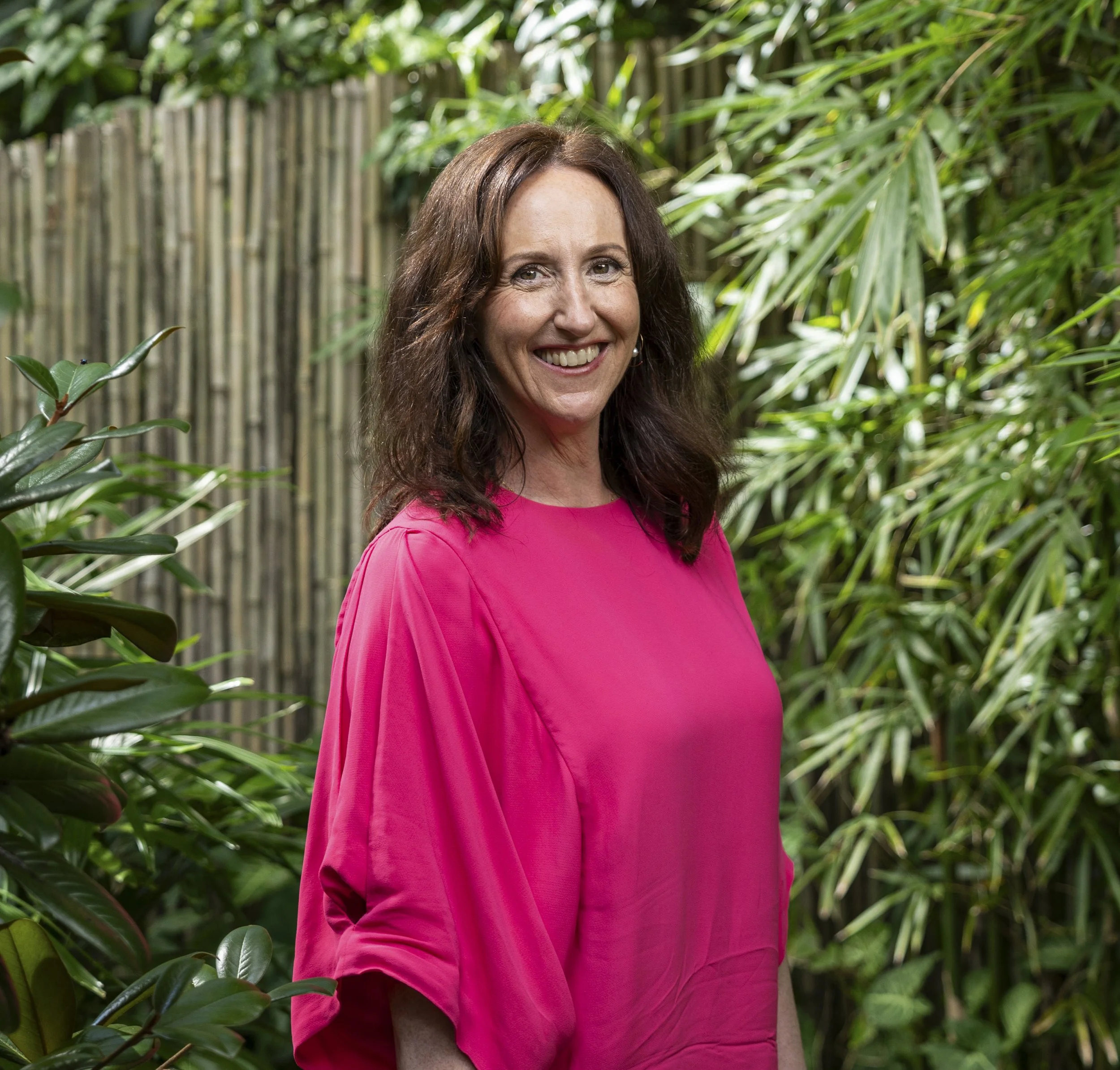 Close-up of a smiling woman with long brown hair, wearing a bright pink top, standing outdoors surrounded by green plants and bamboo fencing.